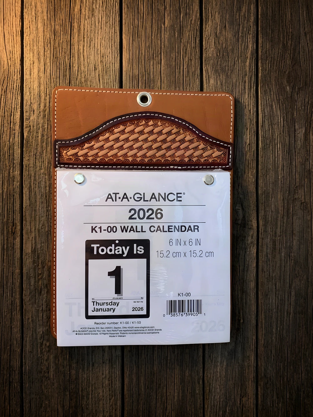 A tooled leather wall calendar holder with a basketweave flap, a hanging grommet and a day-per-page pad, on barn wood.