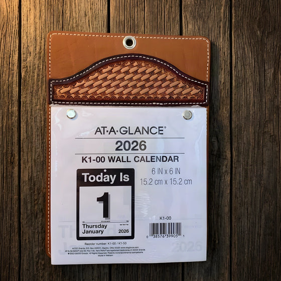 A tooled leather wall calendar holder with a basketweave flap, a hanging grommet and a day-per-page pad, on barn wood.
